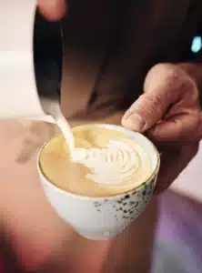 Die Hand eines Barista gießt geschäumte Milch in eine Tasse Kaffee und erzeugt auf der Oberfläche eine komplizierte Latte Art. Die Tasse ist weiß mit dezenten gesprenkelten Mustern. Der Hintergrund ist verschwommen, sodass die Aufmerksamkeit auf den Latte-Art-Prozess gelenkt wird.