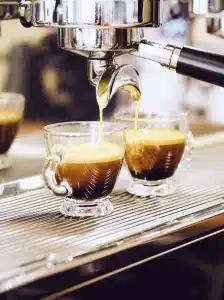Zwei Tassen aus klarem Glas stehen unter einer glänzenden Espressomaschine, in die reichhaltiger, goldener Kaffee fließt und eine Schicht Crema bildet. Der Hintergrund ist verschwommen und lenkt die Aufmerksamkeit auf den Brühvorgang und den verführerischen Kaffee.