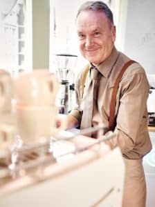 Ein fröhlicher Barista in beigem Hemd, braunen Hosenträgern und Krawatte lächelt hinter einer Theke mit gestapelten weißen Tassen in einem hellen Café. Im Hintergrund ist eine Kaffeemühle zu sehen.