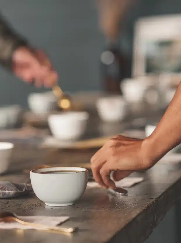 Zwei Personen bereiten an einem Tisch Kaffee zu. Eine Person rührt mit einem Löffel in einer Tasse, die andere streckt ihre Hand nach einer Tasse aus. Mehrere weiße Tassen und Kaffeezubehör sind auf dem Tisch verstreut. Der Fokus liegt auf ihren Händen und den Kaffeetassen.