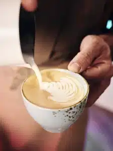 Eine Hand hält eine weiße Keramiktasse mit Kaffee, während eine andere Hand aufgeschäumte Milch hineingießt, wodurch auf der Oberfläche eine kunstvolle Latte Art entsteht. Der Hintergrund ist leicht verschwommen, wodurch der Fokus auf die entstehende Latte Art gelegt wird.