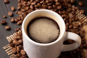 Eine Nahaufnahme einer weißen Keramiktasse mit schwarzem Kaffee, umgeben von verstreuten gerösteten Kaffeebohnen auf einer strukturierten Oberfläche und einem Stück Sackleinen. Der Kaffee hat eine leichte Schaumschicht auf der Oberseite.