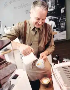 Ein Barista mit grauem Haar und einem Lächeln, der eine beige Schürze und braune Hosenträger über einem hellbraunen Hemd trägt, gießt in einem Café Milch in eine Tasse Kaffee. Eine zweite, fertige Tasse Kaffee mit Latte Art steht neben ihm auf der Theke.