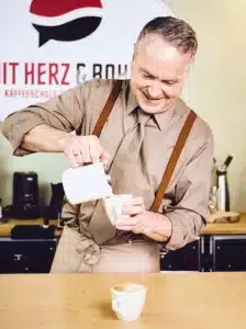 Ein Mann lächelt, während er Milch in eine Tasse Kaffee gießt. Er trägt ein hellbraunes Hemd, eine Krawatte und Hosenträger. Hinter ihm steht ein Tisch mit Kaffeemaschine und eine Wand, an der ein Logo teilweise zu sehen ist.