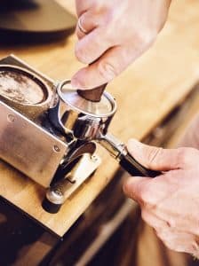 Eine Person presst Kaffeesatz mithilfe eines Tampers in einen Siebträger. Der Siebträger steht auf einer Theke neben einer Kaffeemühle und die Person trägt einen Ring an der linken Hand. Die Szene spielt sich wahrscheinlich in einem Café oder einer privaten Küche ab.