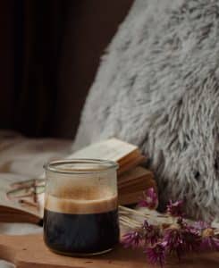 Ein kleines Glasgefäß mit dunklem Kaffee steht auf einer Holzoberfläche. Dahinter sind ein offenes Buch und ein flauschiges graues Kissen zu sehen. Ein Strauß getrockneter lila Blumen liegt neben dem Glas und verleiht der gemütlichen Szene einen Hauch von Farbe.