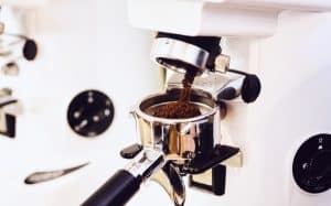Eine Nahaufnahme von gemahlenem Kaffee, der aus einer Kaffeemühle in einen Siebträger gefüllt wird. Der Siebträger befindet sich unter dem Ausguss der Mühle, und der Kaffeesatz fällt hinein. Das Gerät ist vor einem weißen Hintergrund zu sehen.