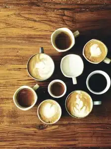 Eine Vielzahl von Kaffeetassen, die auf einem Holztisch angeordnet sind. Die Tassen enthalten verschiedene Kaffeesorten, einige mit aufwendiger Latte Art auf der schaumigen Oberfläche. Der Tisch hat eine reichhaltige, strukturierte Maserung, die zur gemütlichen Atmosphäre der Szene beiträgt.