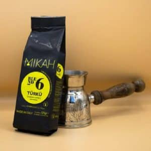 Eine schwarze Tüte türkischen Kaffees/Mocca steht auf einer hellbeigen Oberfläche neben einer traditionellen Cezve-Kaffeekanne aus Metall mit Holzgriff. Die Kaffeetüte hat gelbe Etiketten mit Text. Der Hintergrund ist ebenfalls beige.