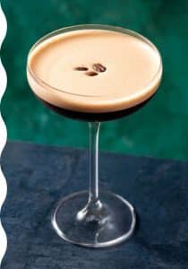 Eine Nahaufnahme eines Espresso-Martini in einem klaren Cocktailglas mit cremigem Schaum obenauf, garniert mit drei Kaffeebohnen. Der Hintergrund hat eine dunkelblaue Oberfläche und einen grünen, strukturierten Hintergrund.