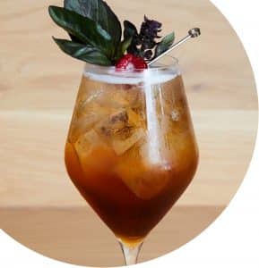 Ein erfrischender Eistee-Cocktail, serviert in einem Weinglas, garniert mit Basilikumblättern und einer Himbeere, gehalten mit einem Cocktailspieß. Das Getränk scheint gut gekühlt zu sein, mit sichtbaren Eiswürfeln und Kondenswasser auf dem Glas.