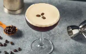 Ein Cocktailglas mit einem Espresso Martini, obenauf eine Schicht Schaum und drei Kaffeebohnen, steht auf einer grauen, strukturierten Oberfläche. Um das Glas herum befinden sich ein Cocktail-Shaker aus Metall, ein Messbecher und verstreute Kaffeebohnen.