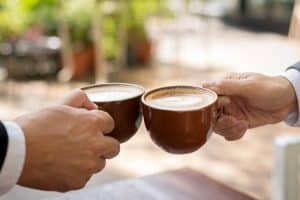 Zwei Hände, die jeweils eine braune Kaffeetasse aus Keramik halten, stoßen zum Anstoßen an. Der Hintergrund ist verschwommen, lässt aber eine Außenkulisse mit grünem Laub und einem Holztisch vermuten. Beide Personen scheinen Geschäftskleidung zu tragen.