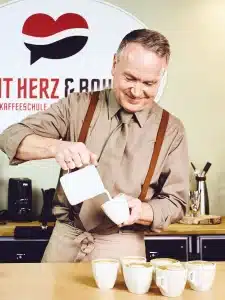 Ein Mann in beigem Hemd und Krawatte sowie Hosenträgern gießt in einem Café Milch in eine Tasse Kaffee. Auf der Theke vor ihm stehen ordentlich mehrere weiße Tassen Kaffee. Hinter ihm ist ein Schild, auf dem ein Teil des Textes zu sehen ist.