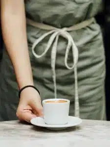 Eine Person mit einer grünen Schürze und einem schwarzen Perlenarmband serviert eine Tasse Kaffee auf einer Untertasse. Die Kaffeetasse steht auf einem Marmortisch. Die Schürze ist an der Taille mit einer weißen Schnur zusammengebunden. Die Person ist nur teilweise sichtbar, der Fokus liegt auf der Hand und dem Kaffee.