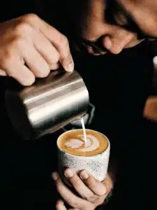 Eine Person gießt gekonnt geschäumte Milch aus einem Metallkrug in eine Tasse Kaffee mit Steinstruktur und erzeugt so ein zartes herzförmiges Latte-Art-Muster auf der Oberfläche des Kaffees. Die Szene ist eine Nahaufnahme, die sich auf die komplizierten Details der Latte Art konzentriert.